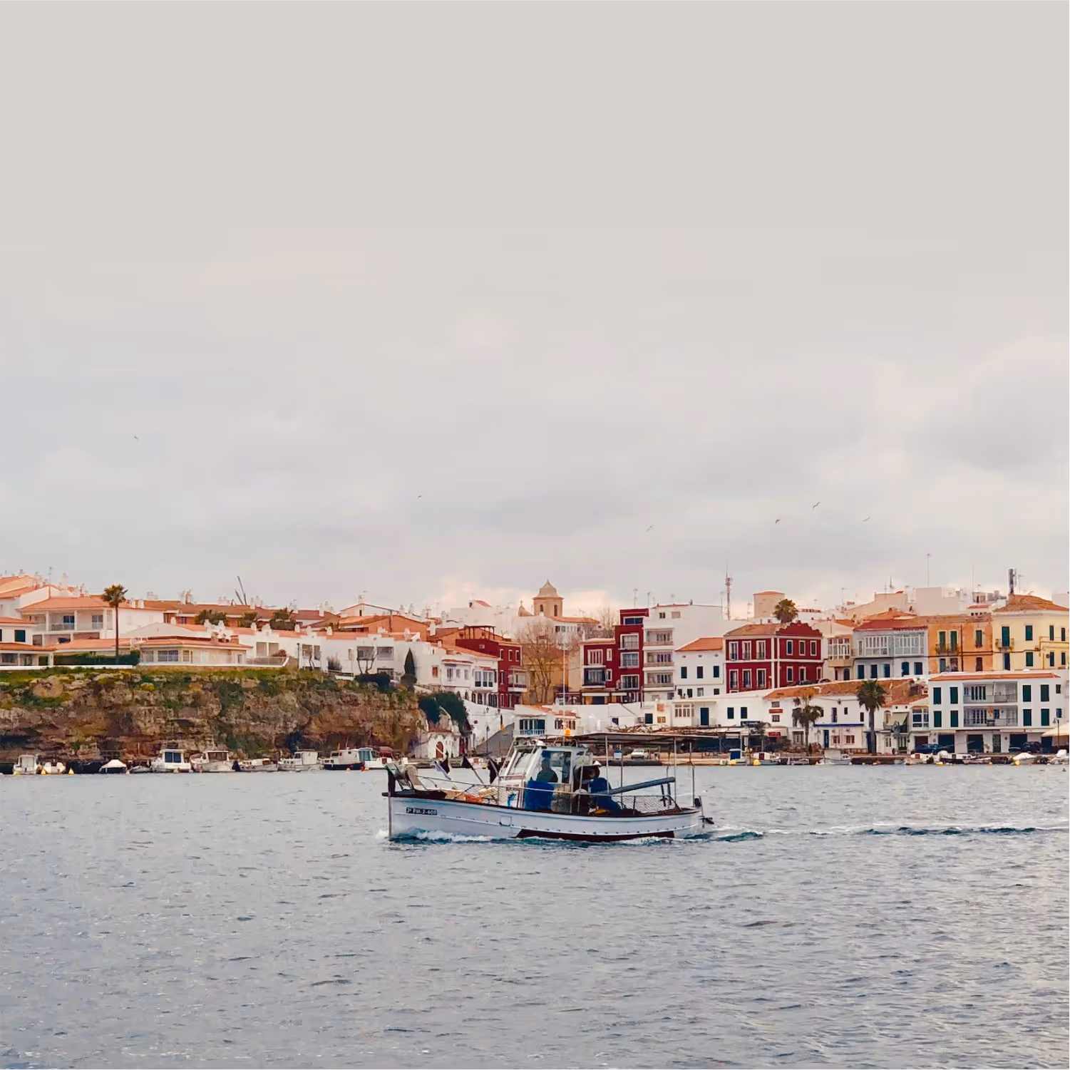 Contact. Image showing a traditional fishing llaut boat sailing through the Port of Mahon. Calesfonts can be seen in the background, with its typical and traditional red and yellow houses that crown the famous fishermen’s harbour in Es Castell.
