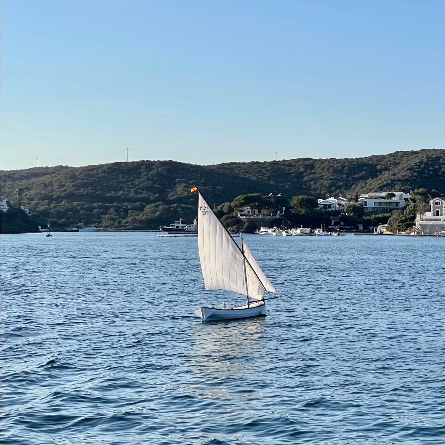 About us, team, partners. Image that shows a traditional latin sail llaut boat sailing through the waters of Port Mahon, with the idyllic shores of Isla del Rey in the background.