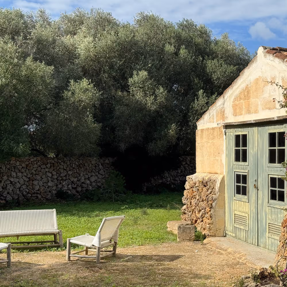 About us, why work with us. Image showing a rustic and rural mares house with tiled roof and a typical green wooden front door. There is a relax setting next to it with sofas and chairs that evoke an idyllic setting for a relaxed conversation. A Typical pared seca wall can be seen in the background, with lush Olive tree canopies above it.