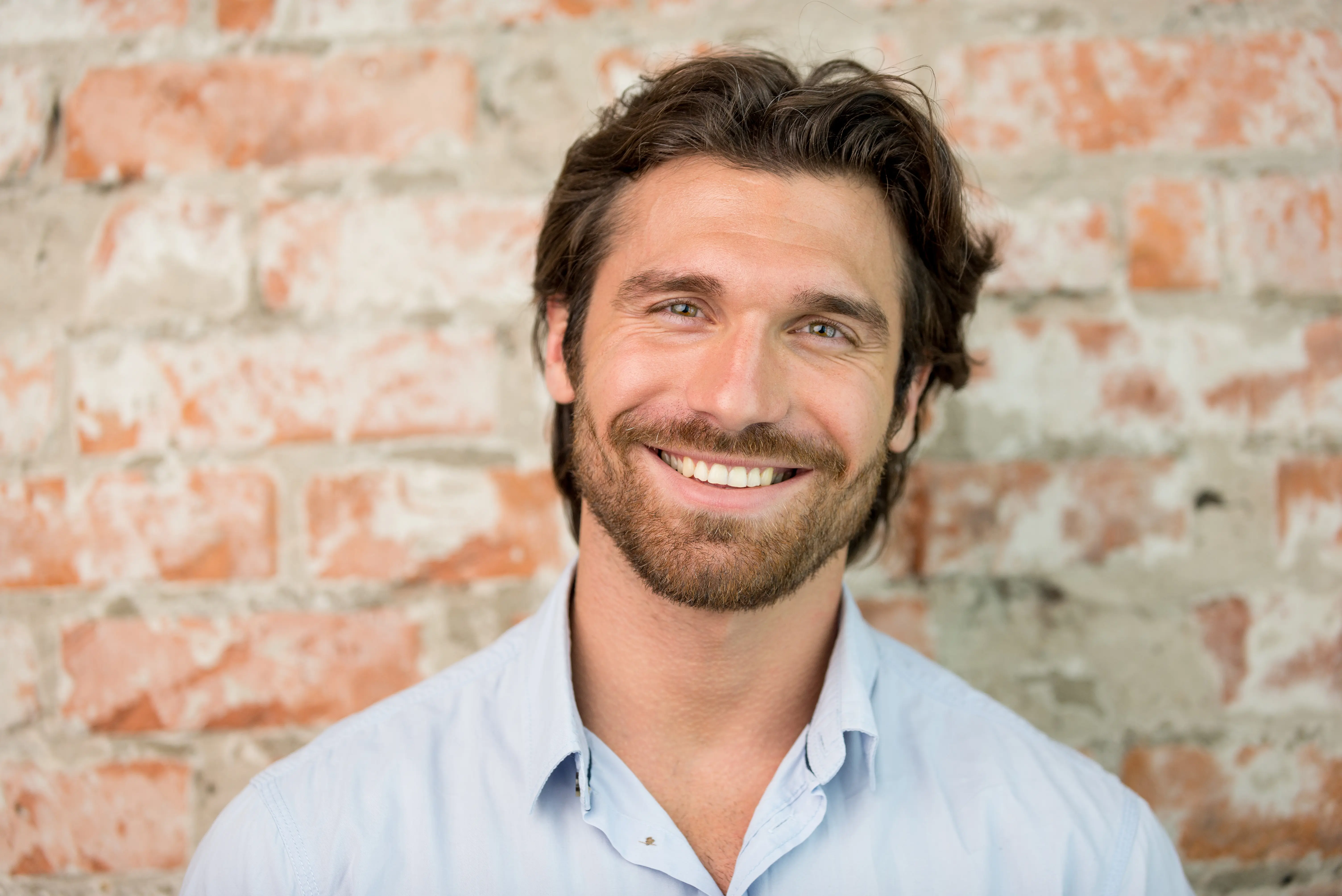 A man with a beard smiling in front of a brick wall.