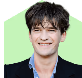 KUGU Mitarbeiter Felix, junger Mann mit zerzausten dunklen Haaren in schwarzem Sakko und blauem Hemd lächelt vor hellgrünem Hintergrund.