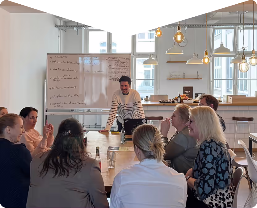 Menschen in einer Besprechung sitzen um einen Tisch, ein Mann steht vor einem Whiteboard und spricht.