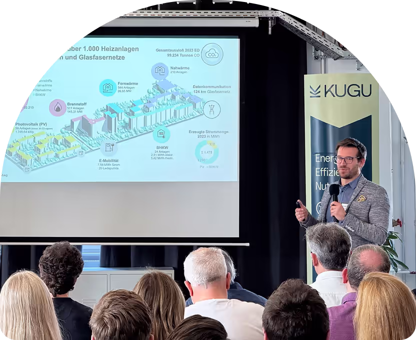 Mann hält einen Vortrag vor Publikum mit einer technischen Präsentation über Heizungsanlagen und Glasfasernetze, neben einem KUGU-Banner.