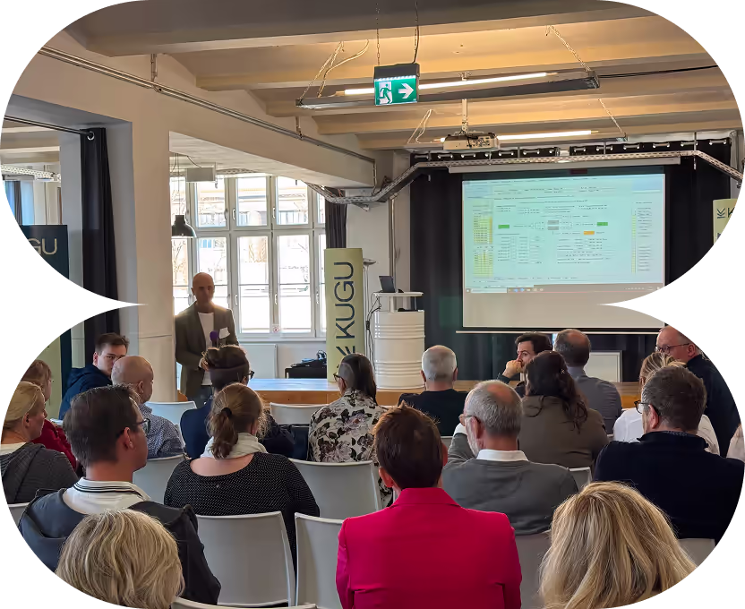 Ein Redner präsentiert vor einem Publikum in einem modernen Raum mit Projektorbildschirm und KUGU-Banner.