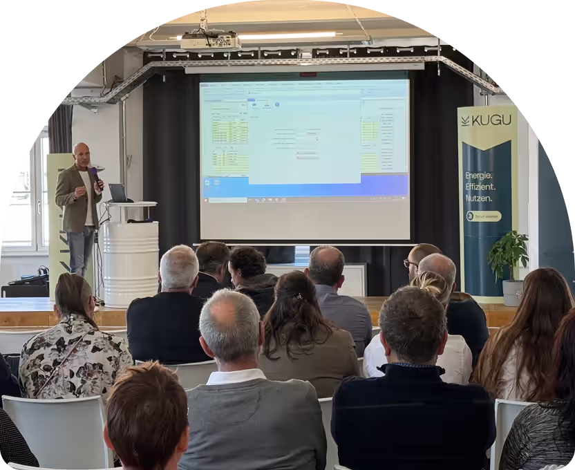 Ein Mann hält eine Präsentation vor Publikum mit einem Bildschirm, der eine Softwareanwendung zeigt, und einem Banner mit der Aufschrift ‚KUGU Energie. Effizient. Nutzen.‘