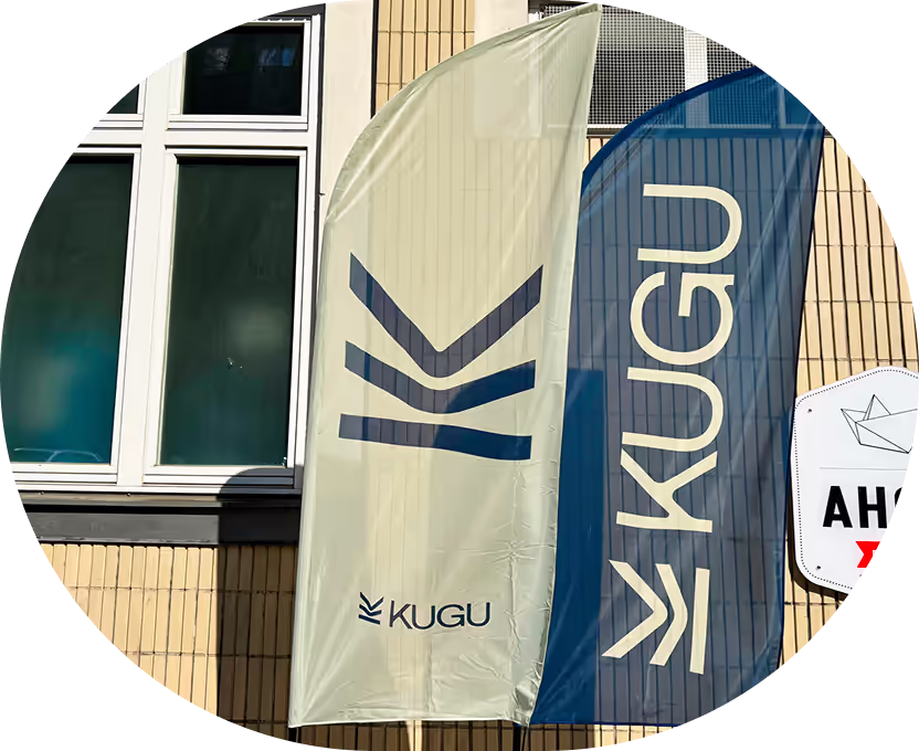 Zwei Fahnen mit dem Logo und Schriftzug KUGU vor einem gebäudefenster.