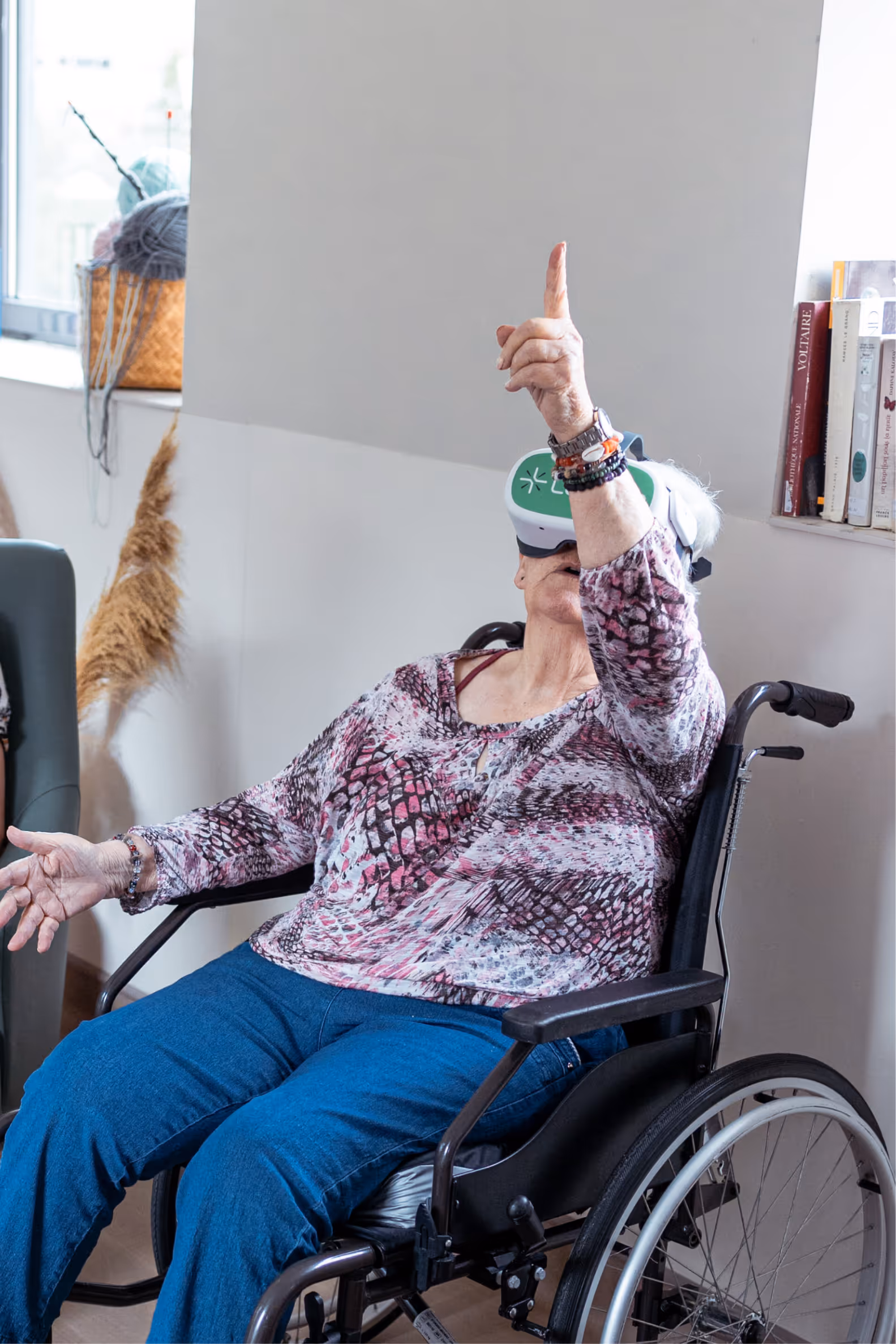 Résidente âgée en fauteuil portant un casque de réalité virtuelle Lumeen, bras levé, en séance immersive apaisante en EHPAD.