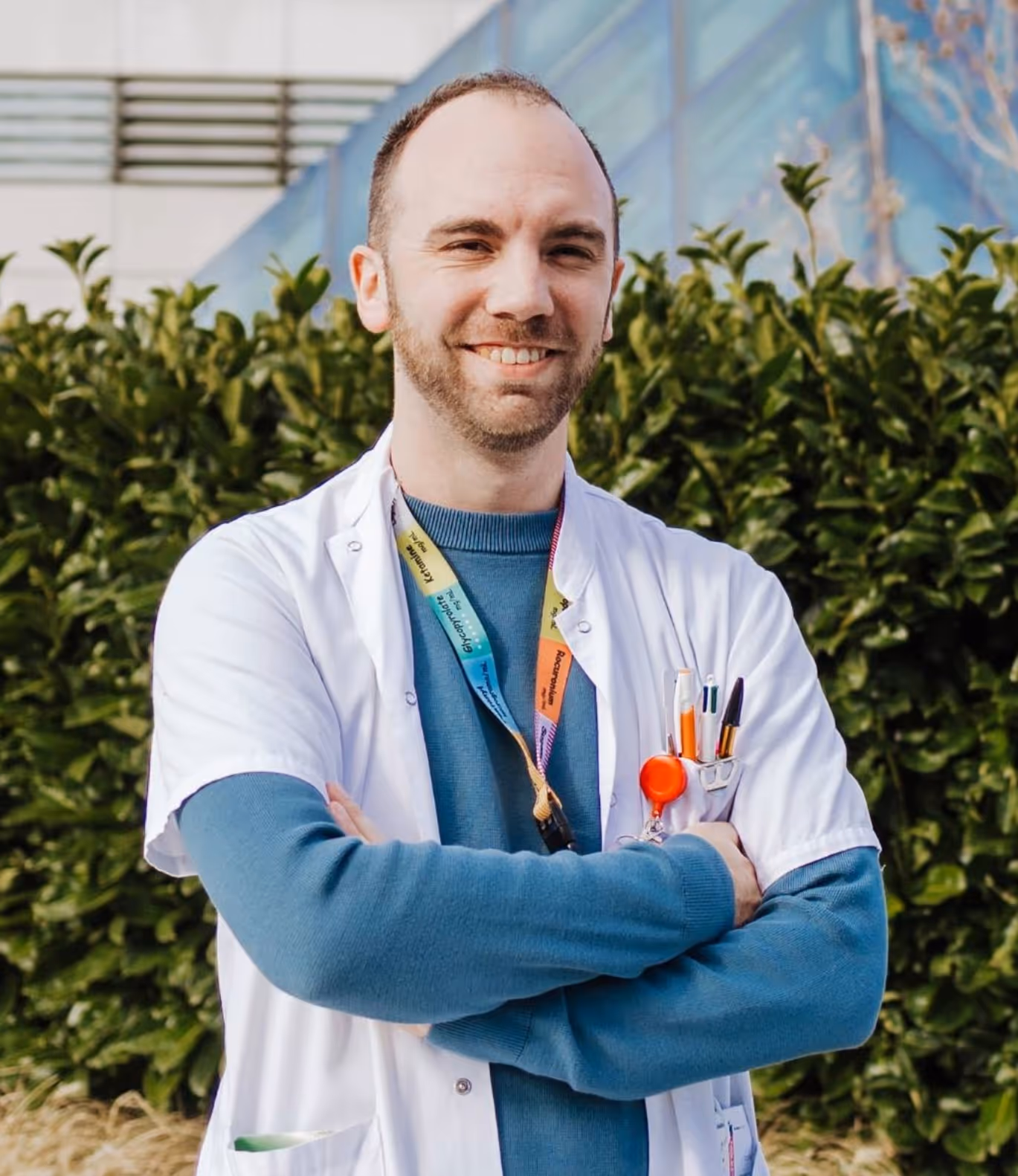 Homme avec une blouse blanche médicale et un polo orange, assis dans un bureau avec un tableau d'affichage et des photos en arrière-plan.
