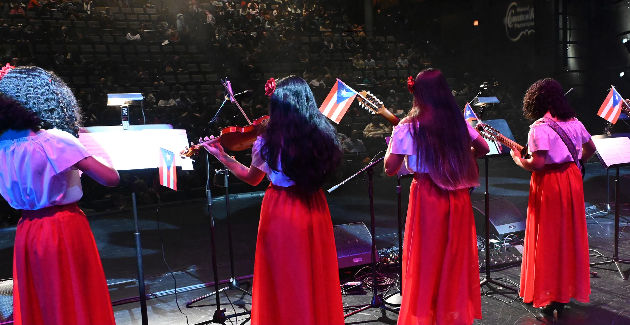 Four female musicians in white tops and long red skirts performing on stage with Puerto Rican flags attached to their music stands.
