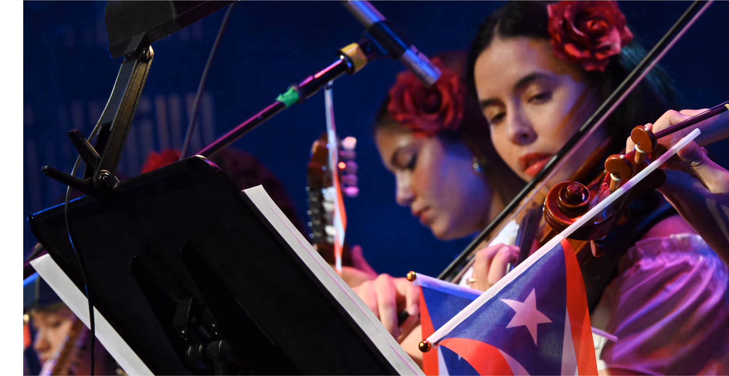 Two female musicians wearing red flowers in their hair playing string instruments with a Puerto Rican flag in front of a music stand.