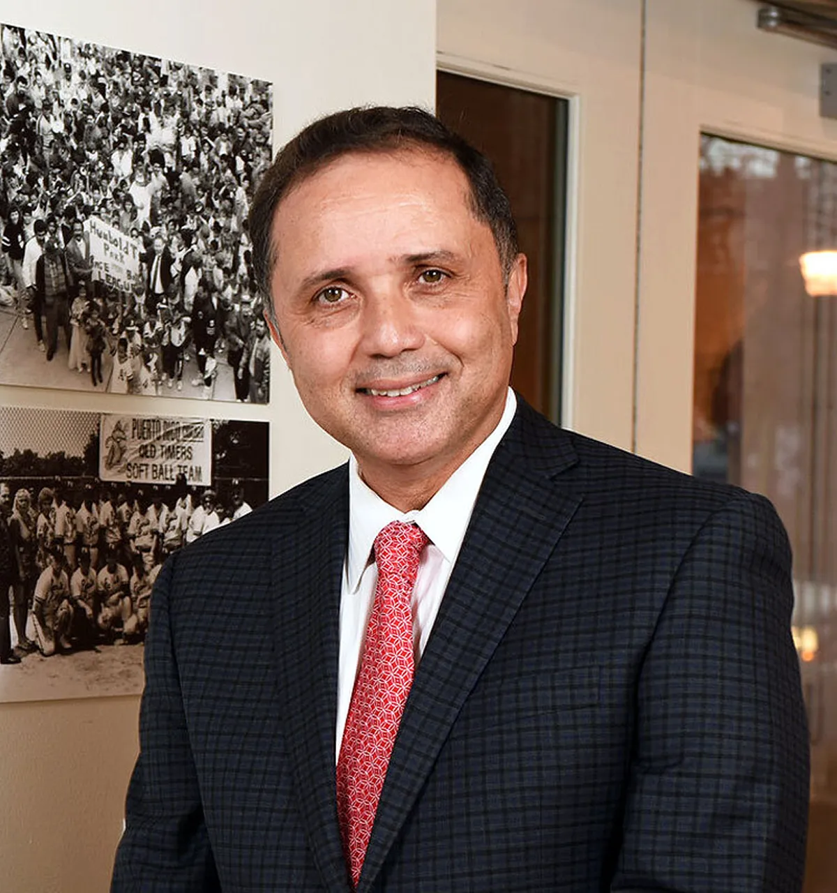 Smiling man in a dark checkered suit and red patterned tie posing indoors with black and white historical photos on the wall behind him.