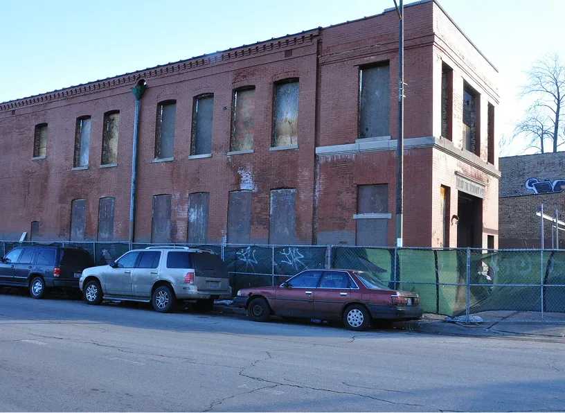 Old two-story brick building with boarded-up windows and a chain-link fence in front.