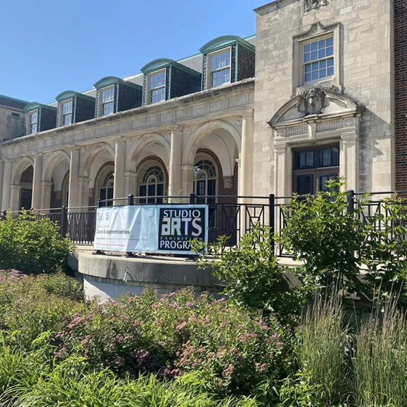 Stone building with arched windows and green dormers, featuring a railing with a banner reading 'Studio Arts Exhibition Program' surrounded by green shrubs under a clear blue sky.