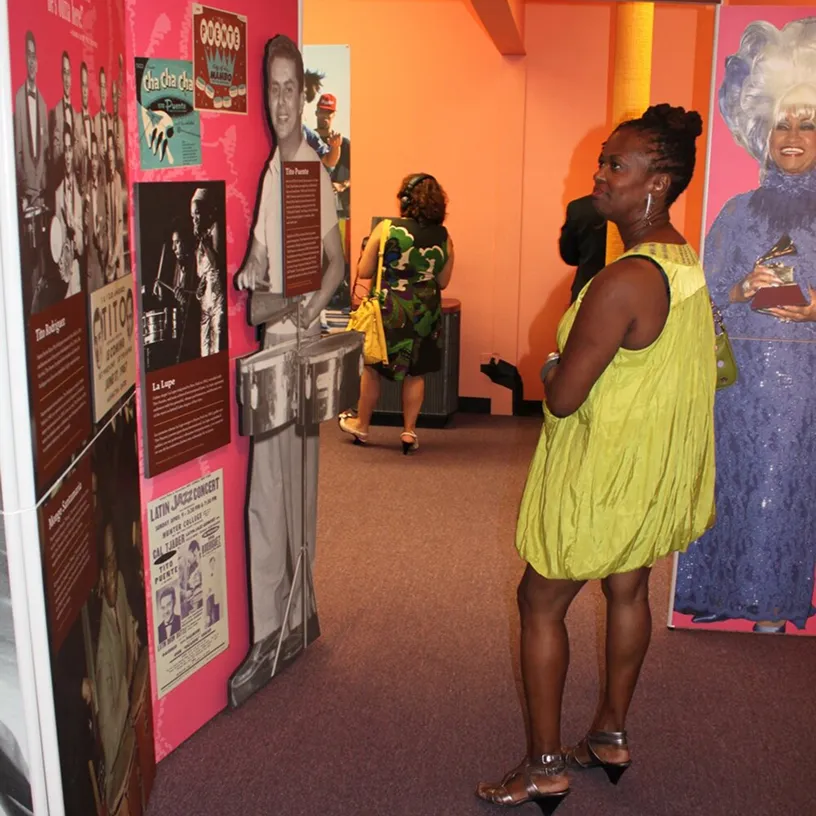Woman in a yellow dress viewing a museum exhibit with vintage photos and a large image of a woman holding a Grammy award.