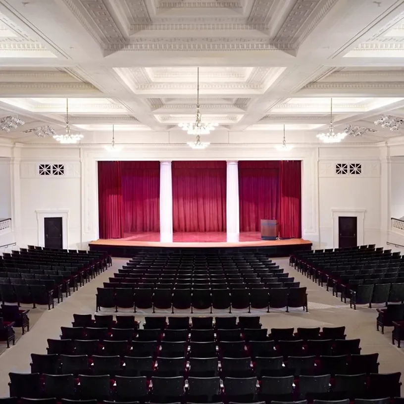 Empty auditorium with rows of black seats facing a stage with red curtains and two white columns.