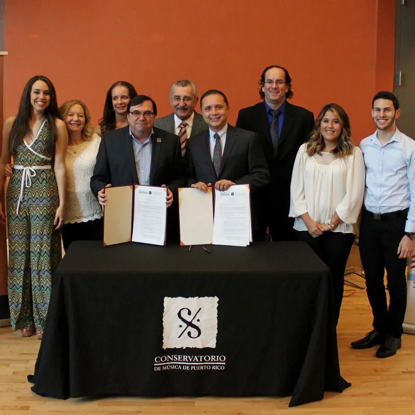 Group of nine people standing behind a table with a black cloth that has the Conservatorio de Música de Puerto Rico logo, two men in the front holding open documents.