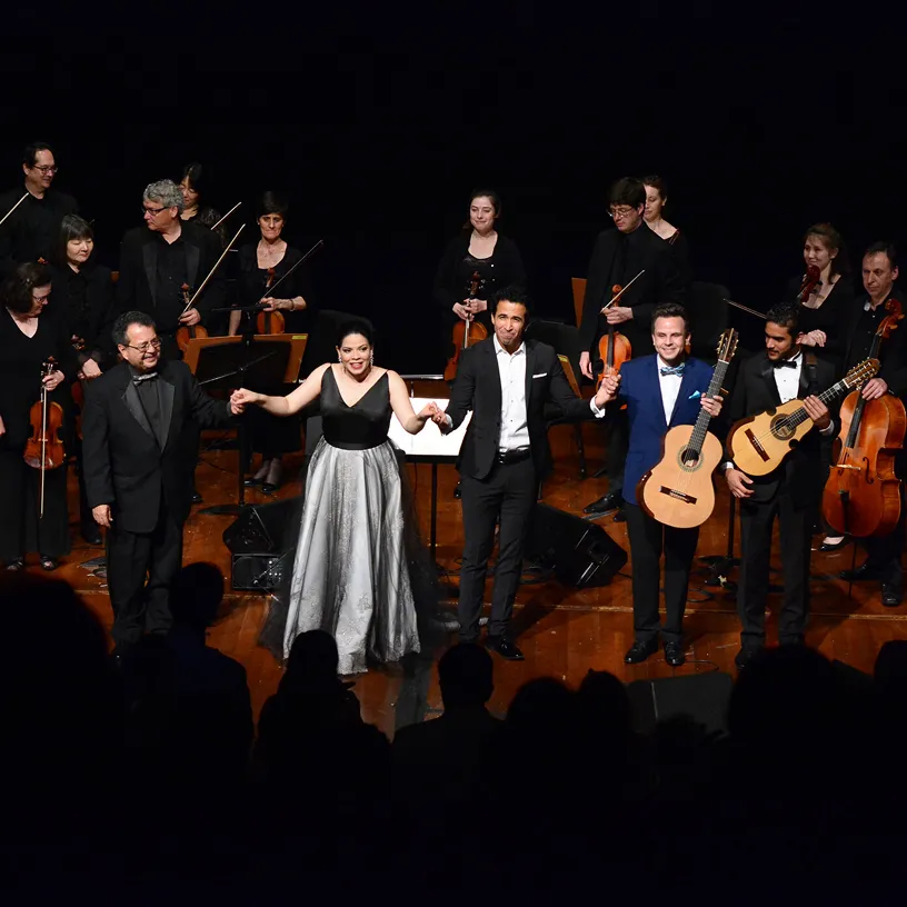 Group of musicians and performers holding hands and taking a bow on stage after a concert, with string orchestra in the background.