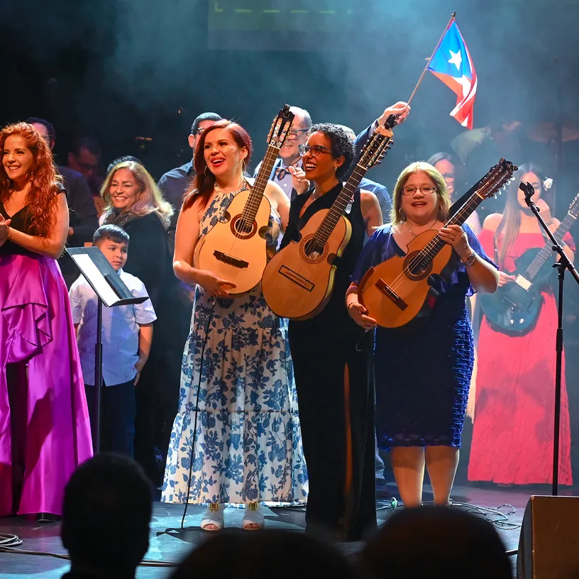 Three women on stage holding guitars, with one man in the background waving a Puerto Rican flag during a musical performance.