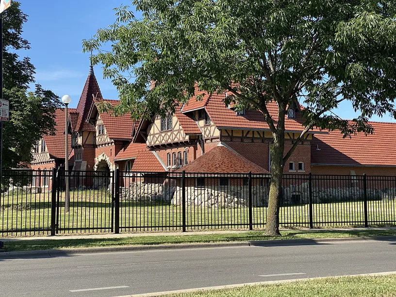 Large Tudor-style building with red tiled roofs behind a black metal fence and a tree in the foreground on a sunny day.
