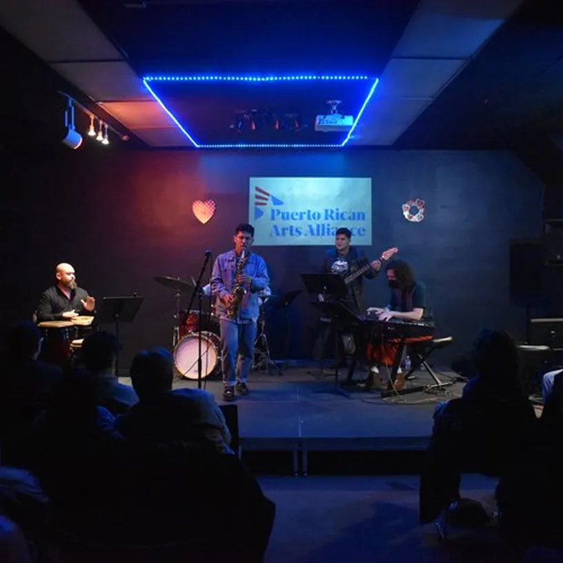 Musicians performing on stage with saxophone, keyboard, bass guitar, and percussion under blue neon lights and a Puerto Rican Arts Alliance sign.
