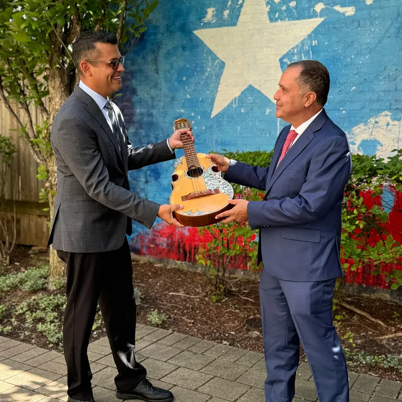 Two men in suits smiling and exchanging a small acoustic guitar outdoors in front of a painted wall with a large white star on blue and red background.