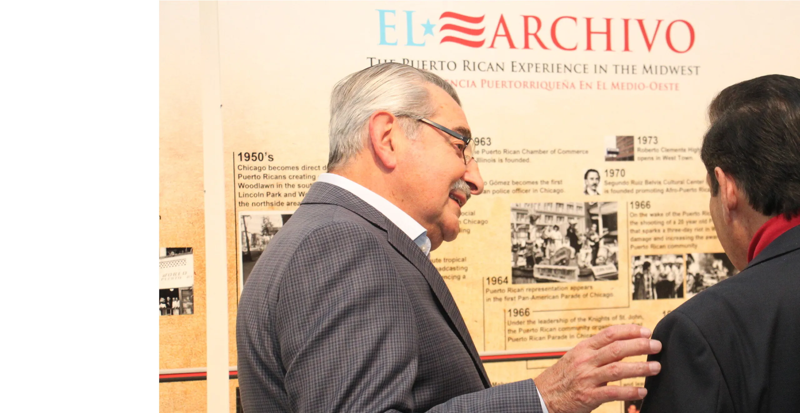 Older man with gray hair and glasses speaking to another man in front of a historical exhibit about the Puerto Rican experience in the Midwest.