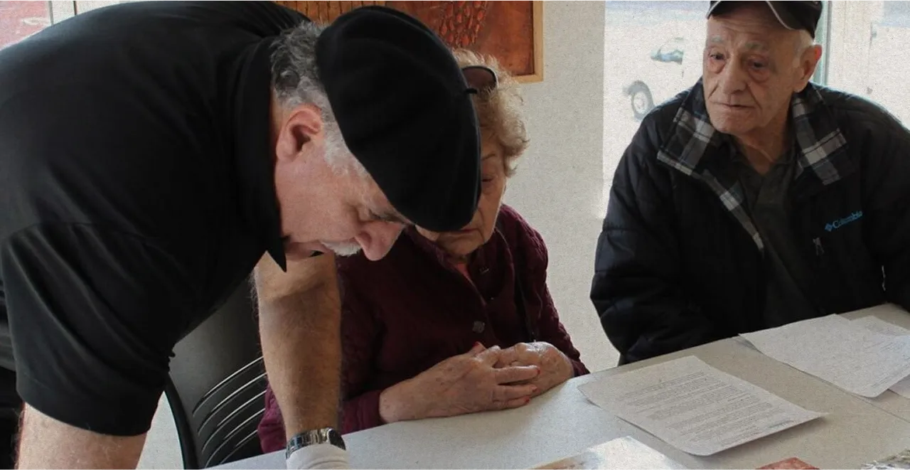 Three elderly people at a table reviewing documents, one man wearing a black beret leans forward.