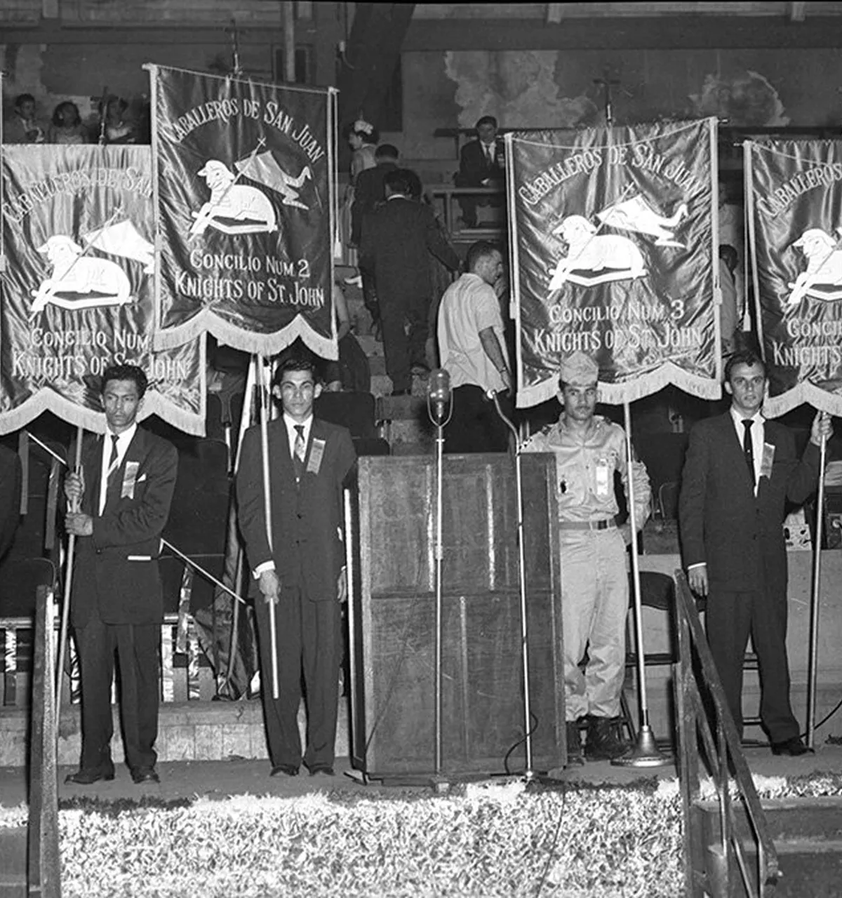 Four men in formal attire and a military uniform stand holding banners labeled 'Caballeros de San Juan, Concilio Num 2 and 3, Knights of St. John' behind a wooden podium with a microphone.