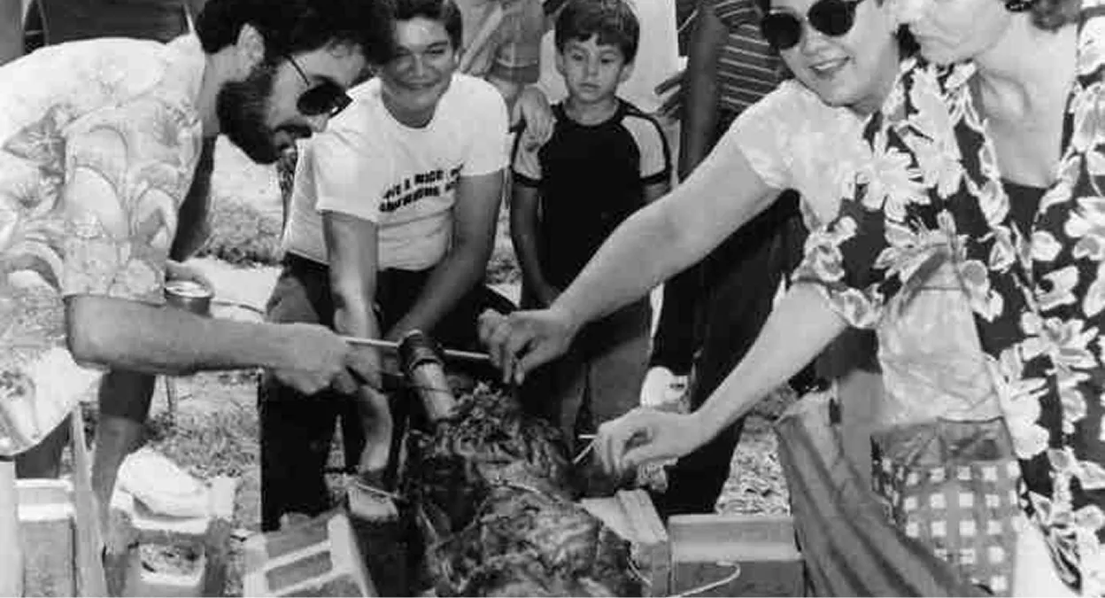 Group of people gathered around a roasting pig at an outdoor event, some wearing sunglasses and patterned shirts.