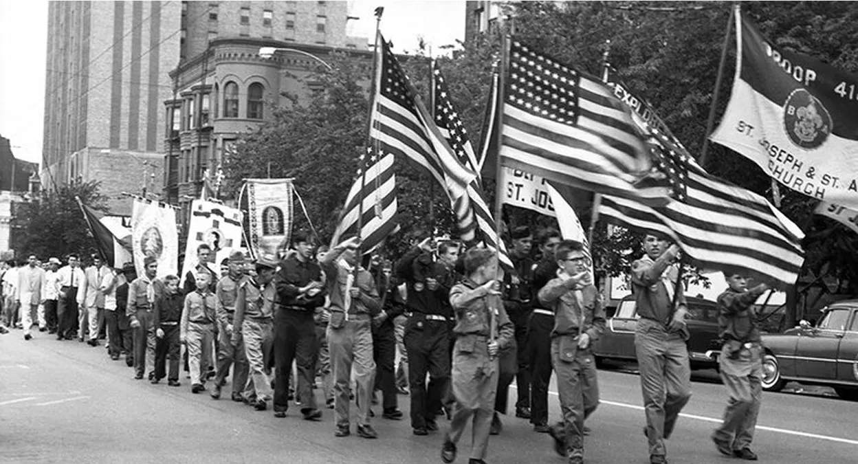 Black and white photo of boys and men marching in a parade, carrying American flags and banners on a city street.