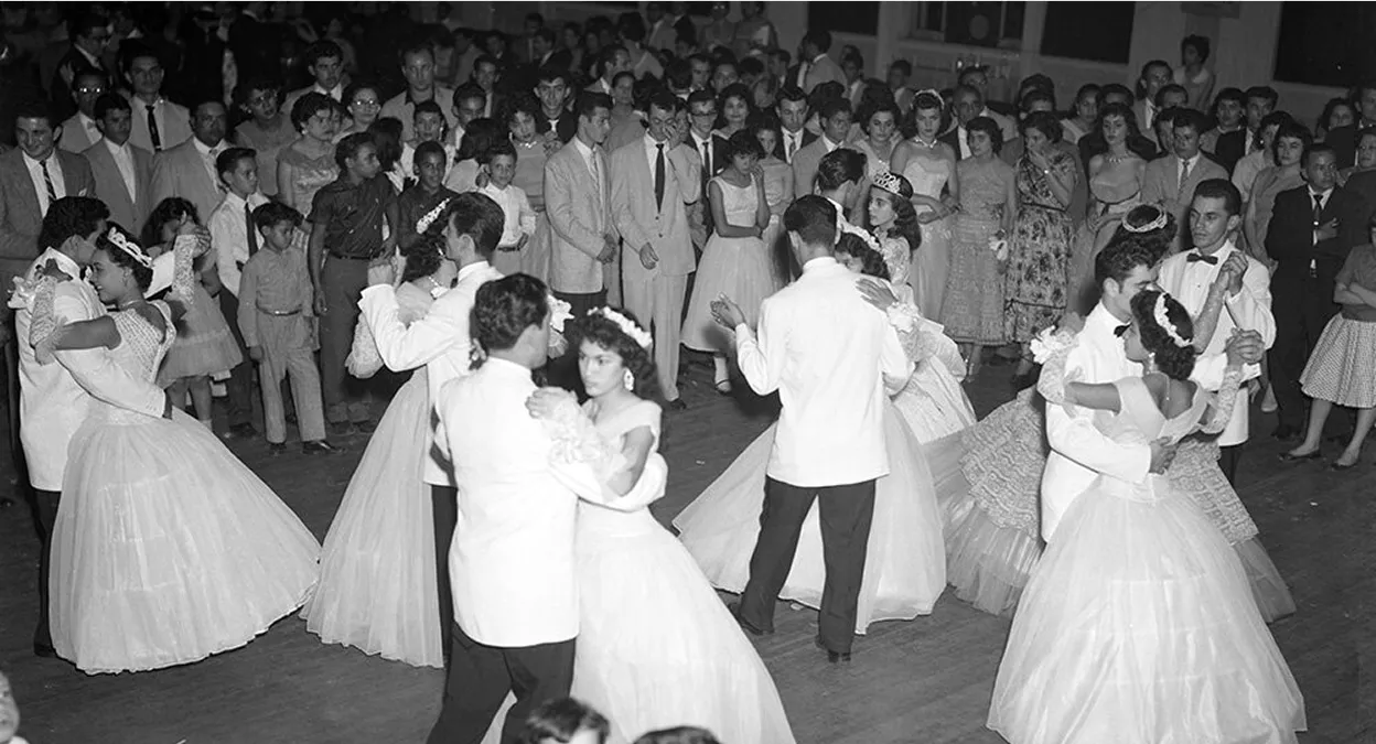 Black and white photo of young women in formal gowns and tiaras dancing with men in white jackets, watched by a crowd.