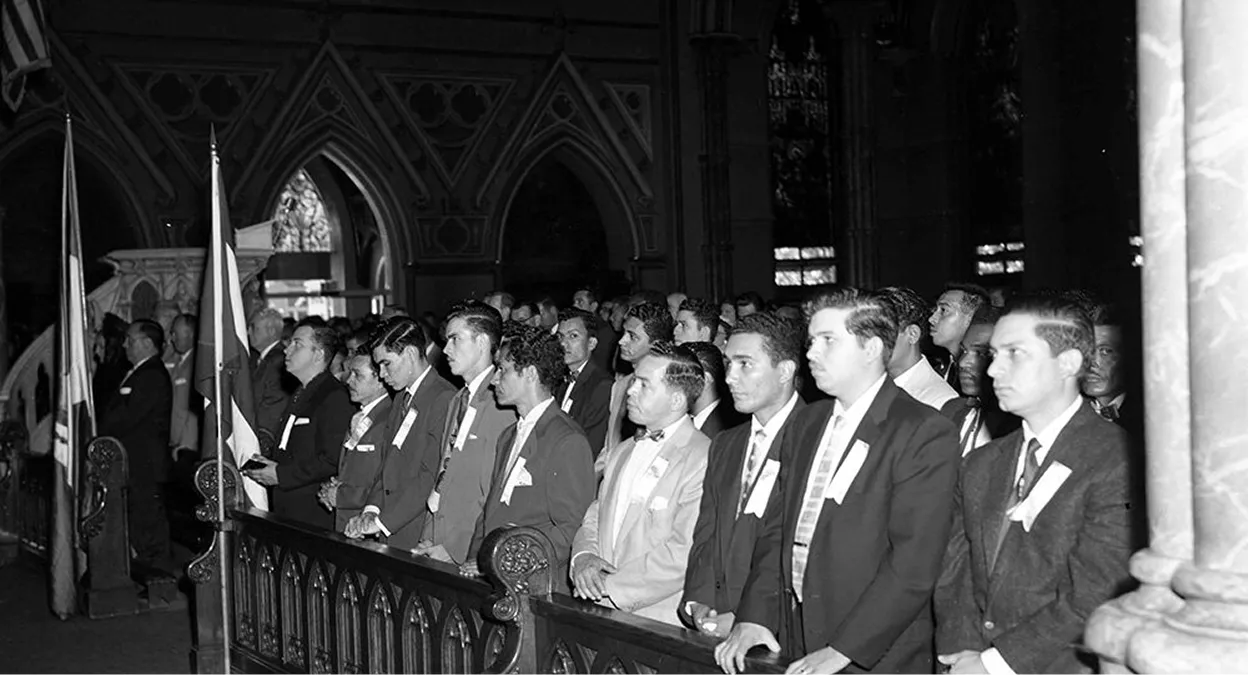 Black and white photo of men in suits standing in rows inside a church with stained glass windows and flags.