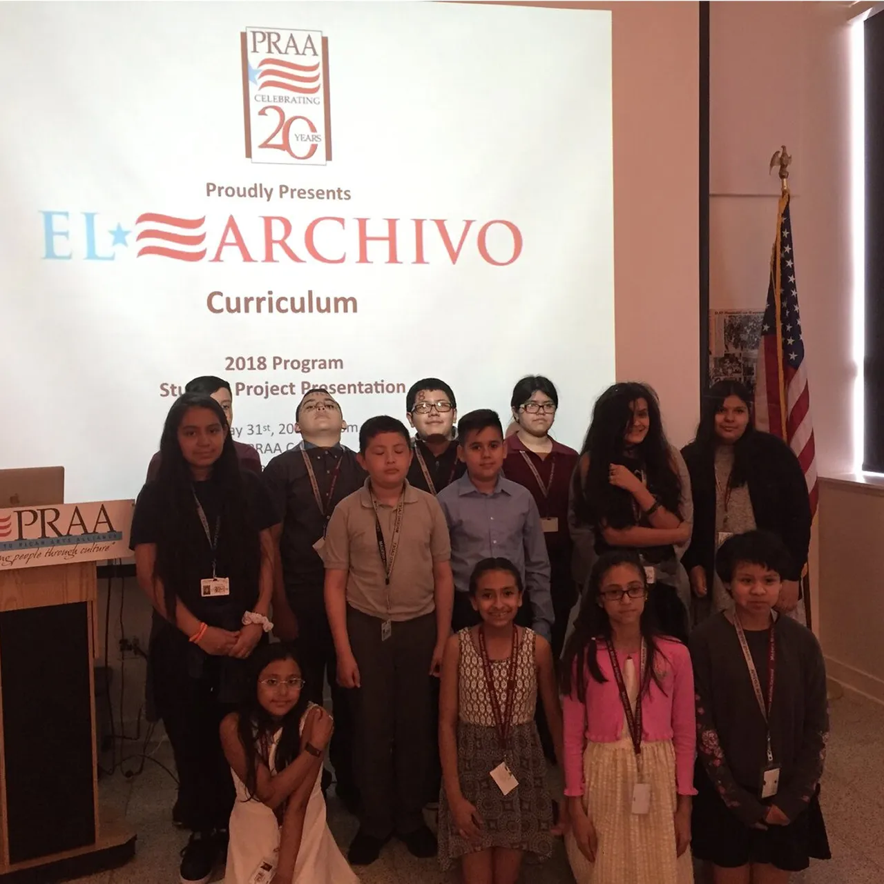 Group of children posing in front of a presentation screen displaying 'EL ARCHIVO Curriculum 2018 Program Student Project Presentation' with a PRAA logo and an American flag in the background.