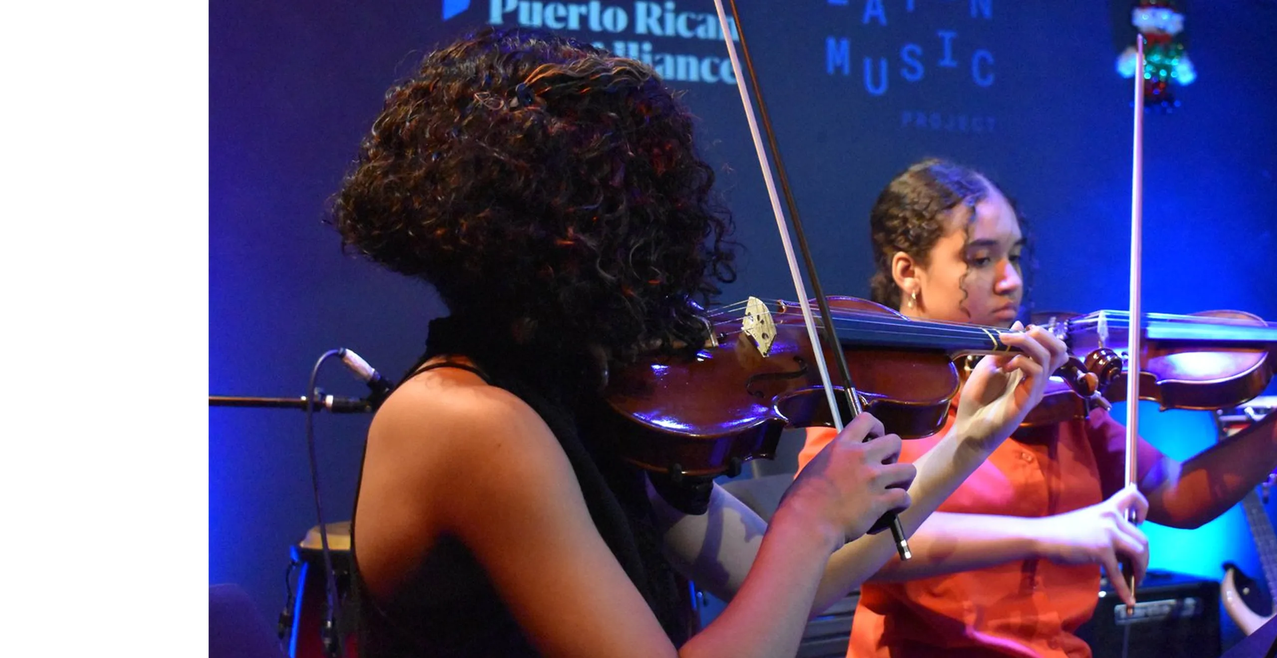 Two young women playing violins on stage with a blue background displaying Puerto Rican Arts Alliance and Latin Music Project text.
