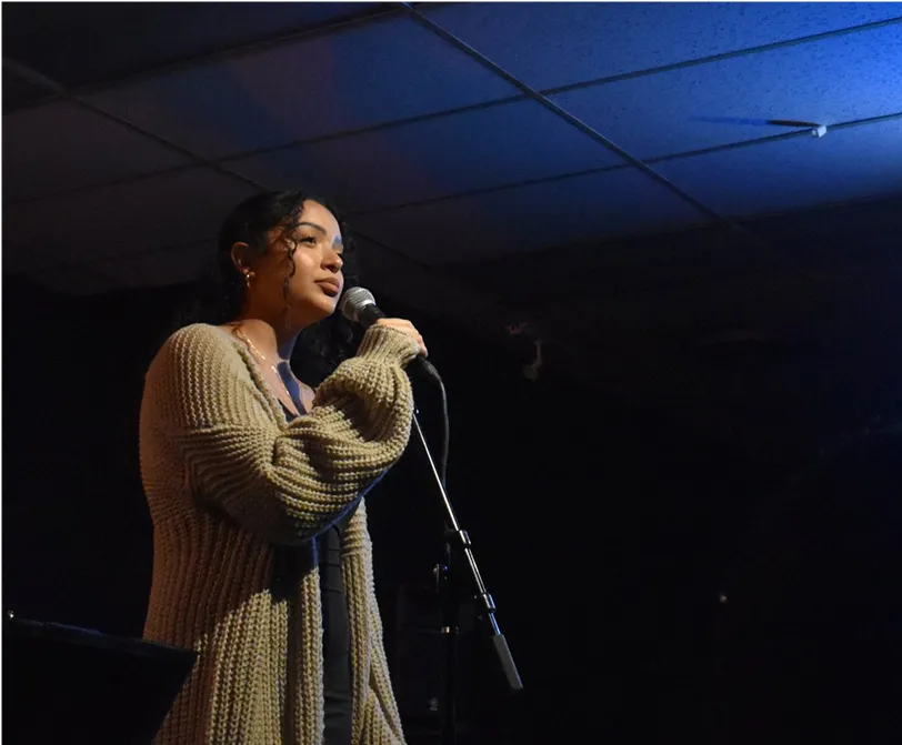 Young woman with curly hair singing into a microphone on stage.