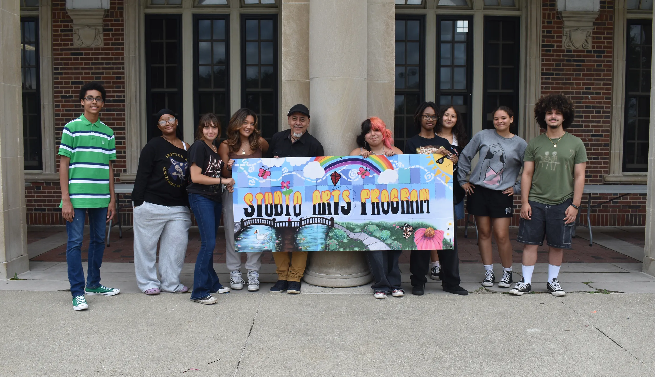 A diverse group of nine young people standing outside a building, holding a colorful banner that reads 'Studio Arts Program.'