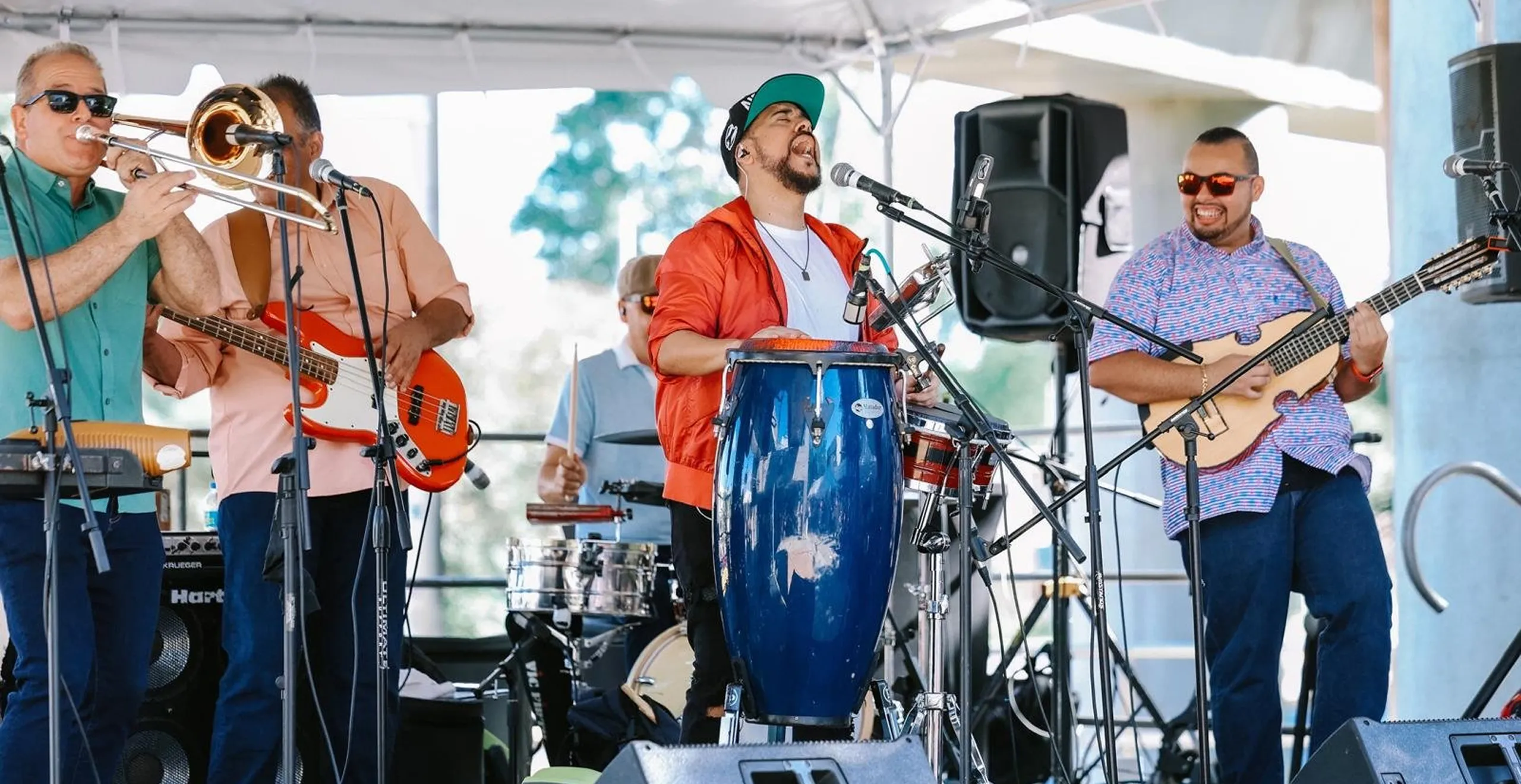 Five-member band performs on stage with trombone, bass guitar, drums, congas, and strings, all wearing colorful clothes and sunglasses.