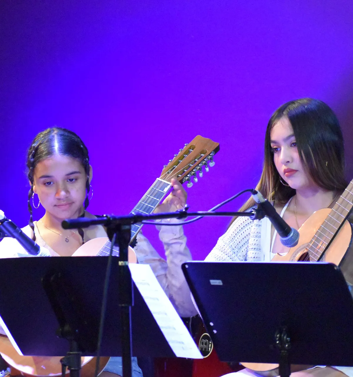 Two young women playing acoustic guitars on stage with music stands and microphones in front of a purple backdrop.