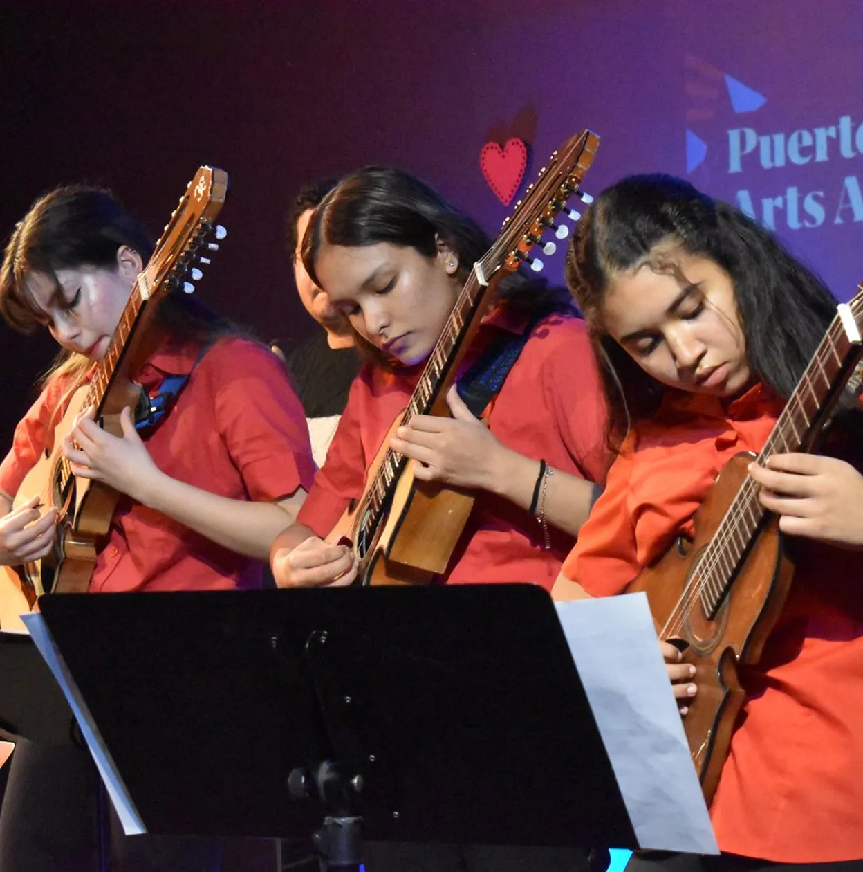 Three young women wearing red shirts playing acoustic guitars on stage with a music stand and sheet in front.