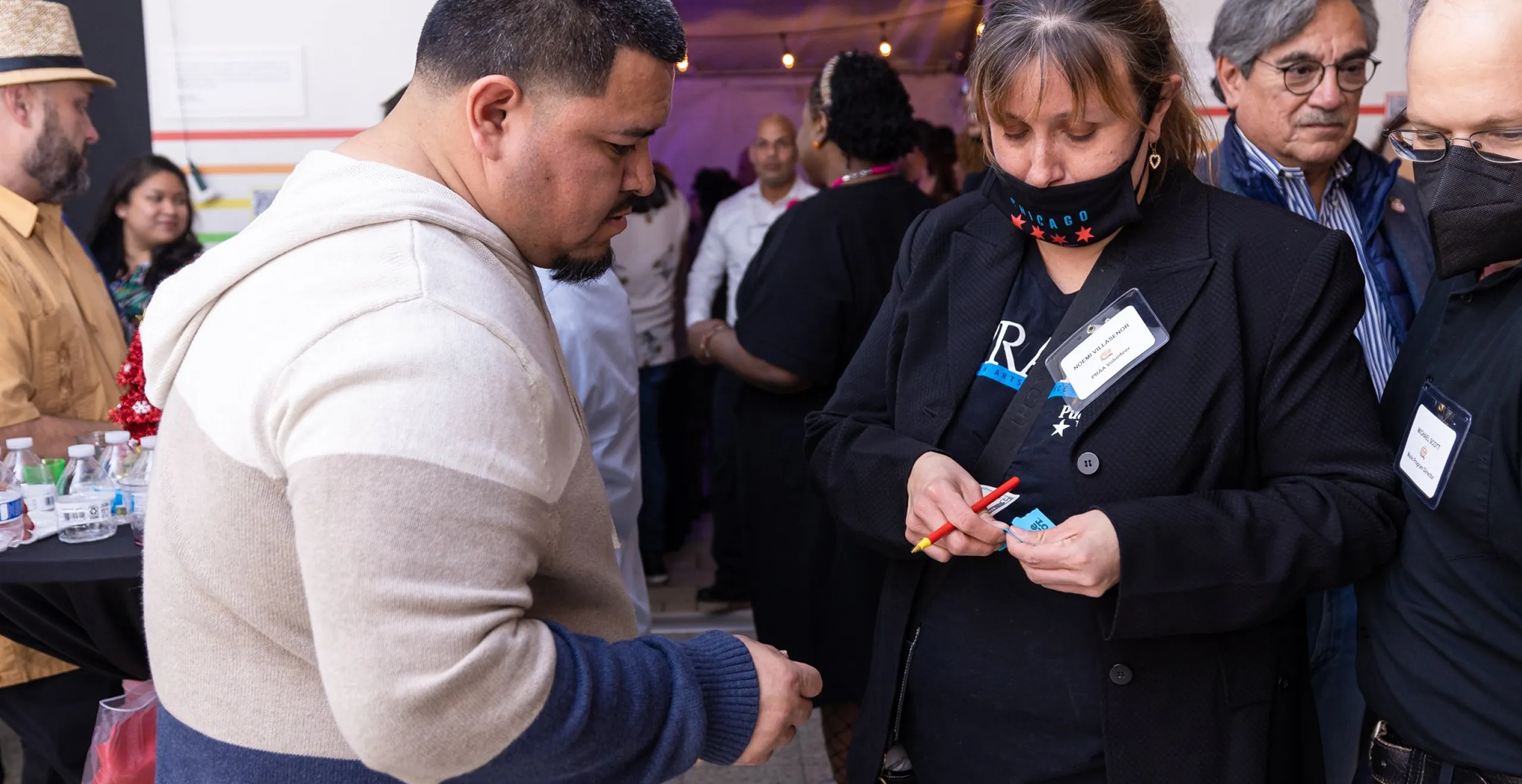 A woman wearing a Chicago face mask and a name tag writes on a blue ticket while a man in a beige and blue hoodie watches; several people are gathered in the background.