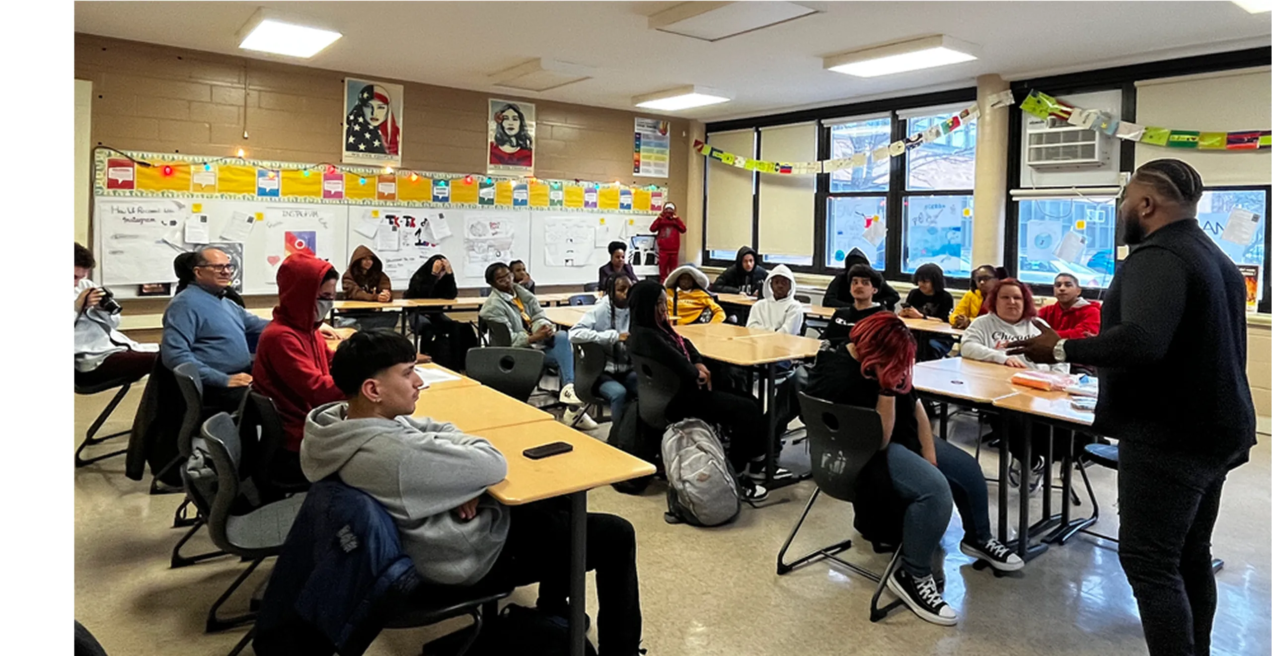 A classroom with students seated at tables listening to a man speaking near the front of the room.