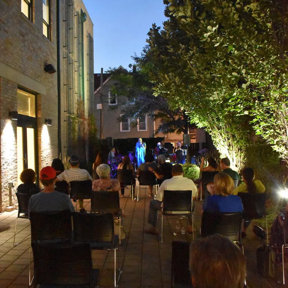 Outdoor evening event with seated audience watching a live musical performance under trees and building lights.