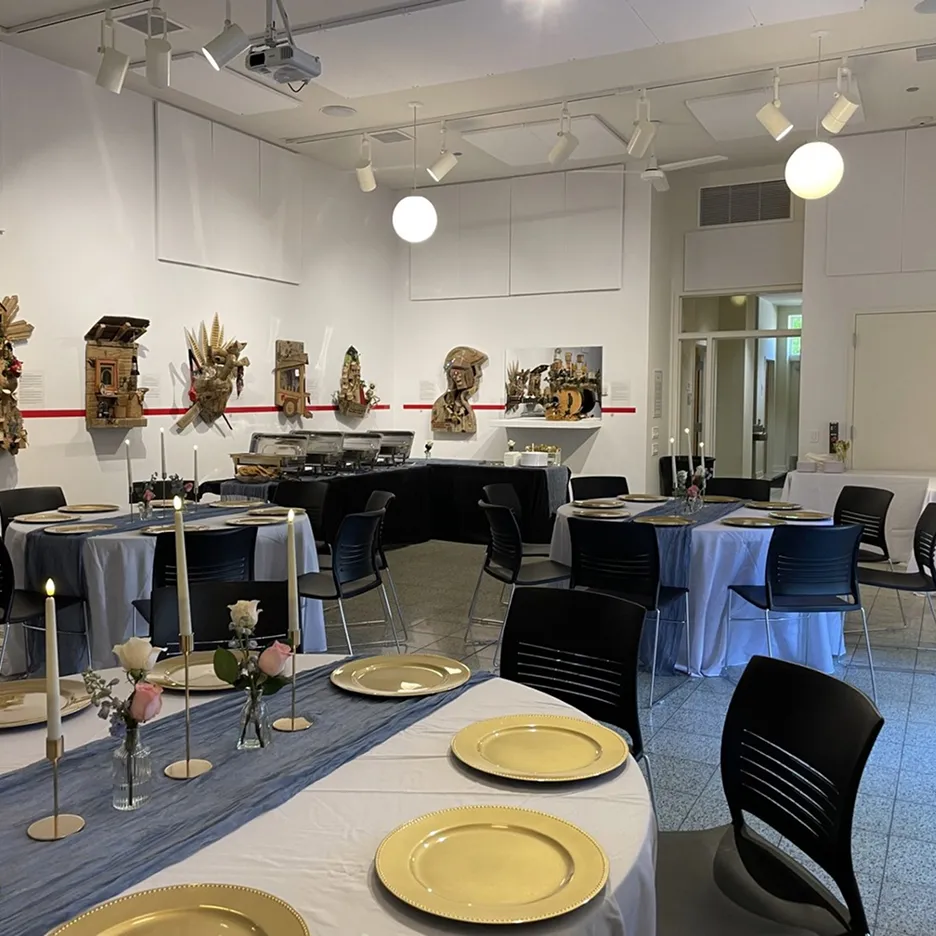 Indoor dining space with round tables set with yellow plates, candles, and small flower arrangements, against a wall displaying artistic sculptures.