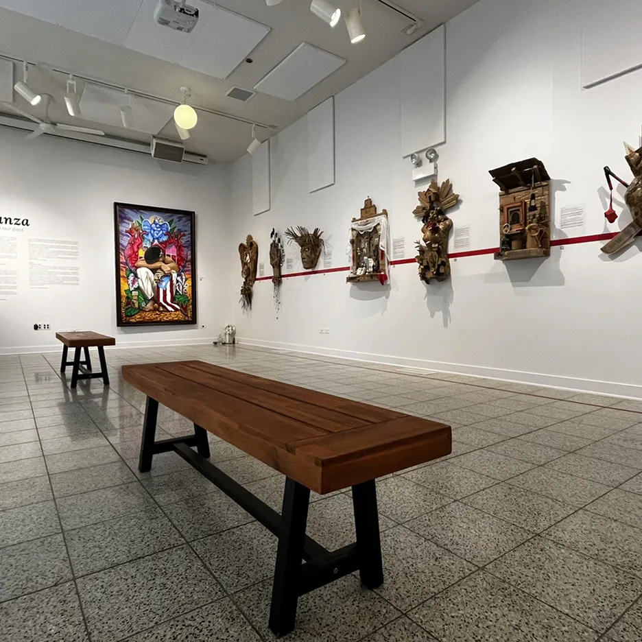 Art gallery interior with wooden benches and wall-mounted mixed-media sculptures and a vibrant framed painting.