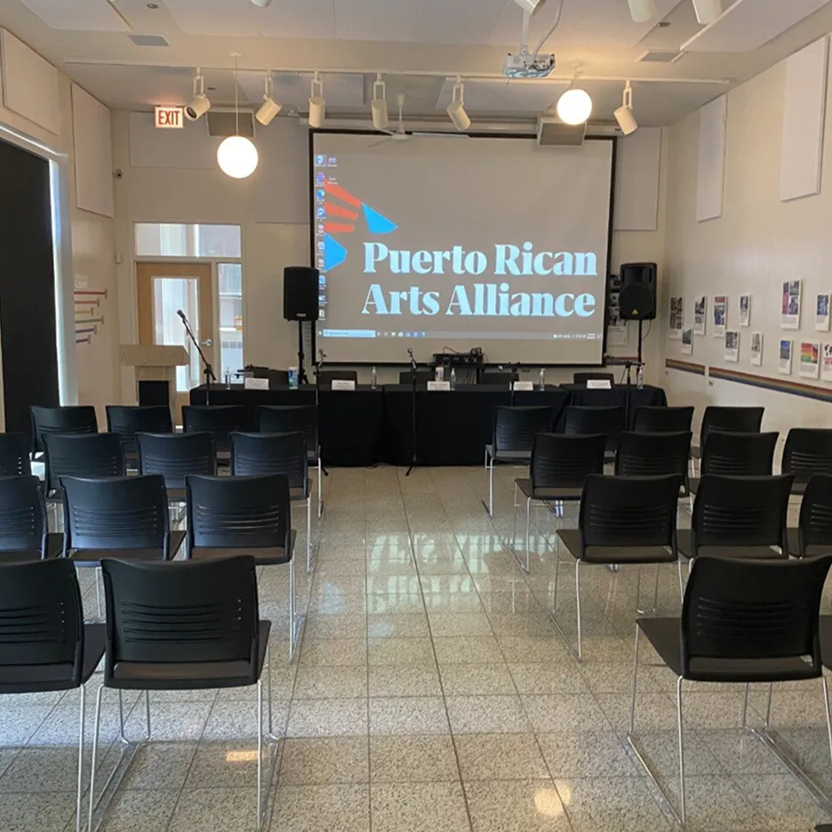 Conference room with rows of black chairs facing a stage and projector screen displaying 'Puerto Rican Arts Alliance'.
