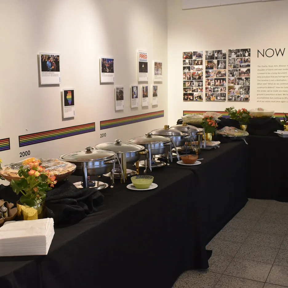 Buffet table with covered chafing dishes, bowls of sauces, flower arrangements, and napkins set in a gallery with a timeline and photos on the wall.