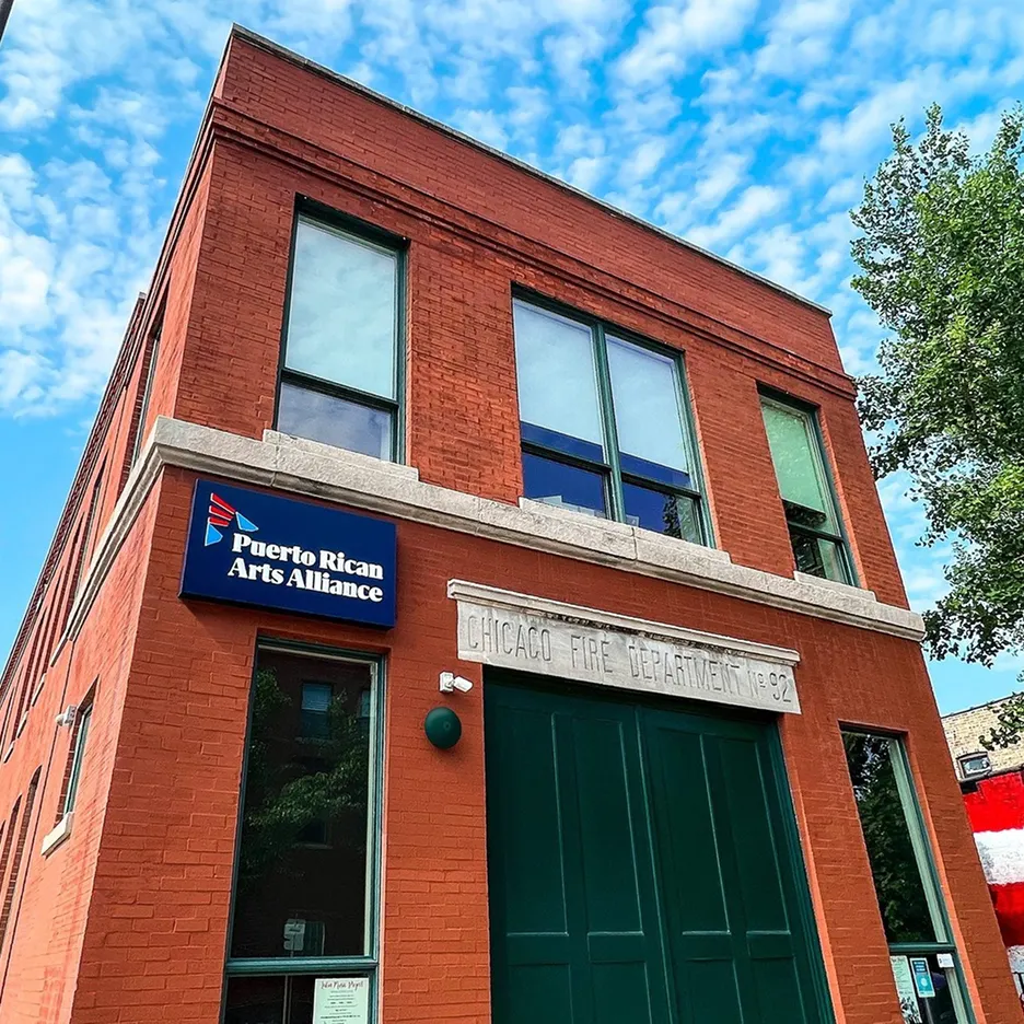 Red brick building with a blue sign reading 'Puerto Rican Arts Alliance' and a stone plaque labeled 'Chicago Fire Department No. 92' under a partly cloudy sky.
