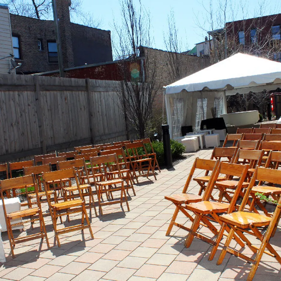 Outdoor patio setup with rows of wooden folding chairs facing a white canopy tent on a paved surface.