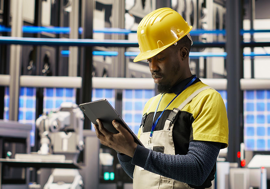 A man wearing a yellow hard hat is looking at a tablet.
