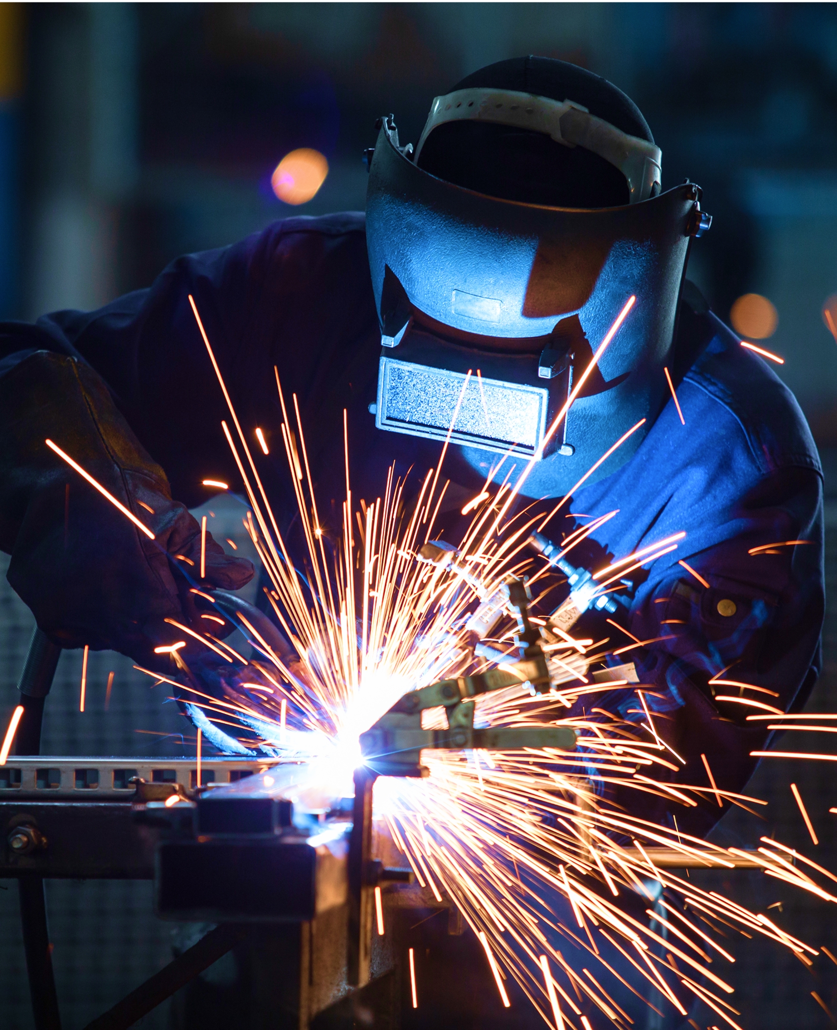 A man wearing a welding helmet and blue coveralls is welding metal.